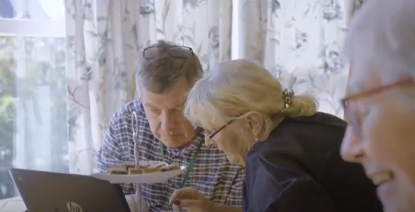 Older man looking at laptop with lady in home setting, and older woman in foreground