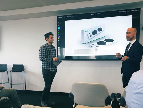 Chris Hughes presenting with Hector Minto of Microsoft in front of a screen showing the XBox adaptive controller