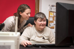 Image shows a woman at a computer. Someone is standing behind them resting a supportive hand on their shoulder