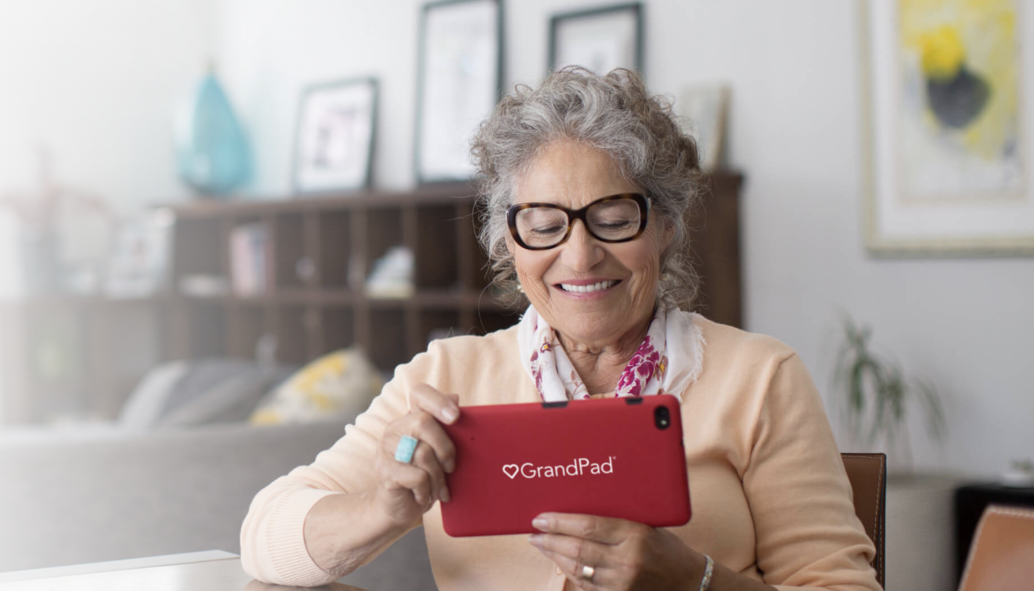 A picture of an older lady cradling the GrandPad tablet. She wears glasses. The tablet is red.