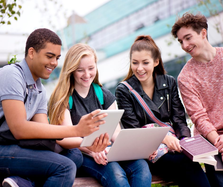 Group of four young people at down outside a college, smiling looking at a tablet and a laptop