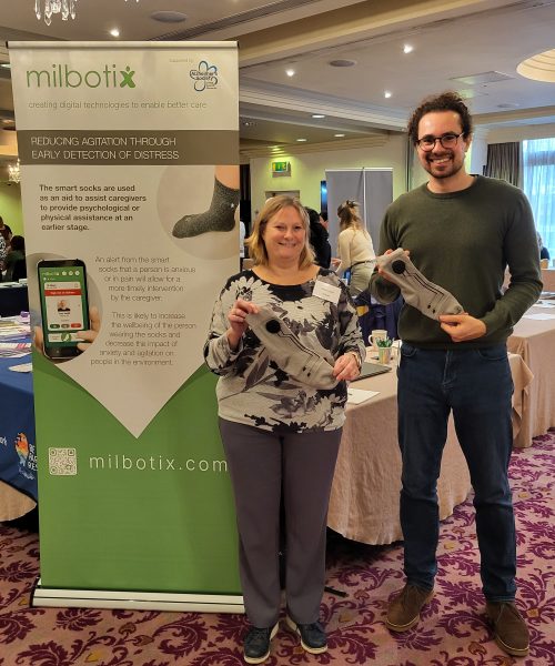Two people stand holding prototypes of SmartSocks in front of a Milbotix banner