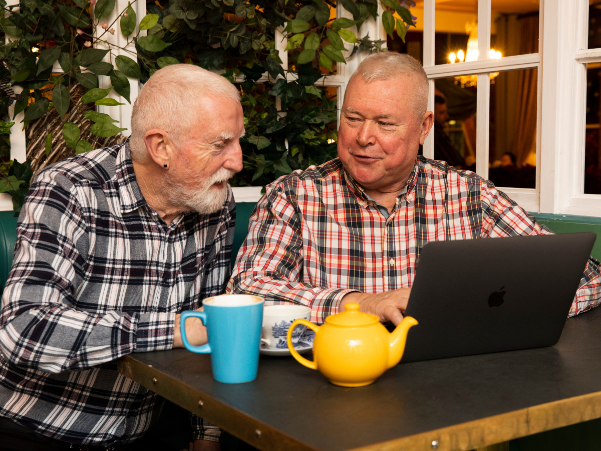 Two older people sitting together in front of a laptop, enjoying a pot of tea.