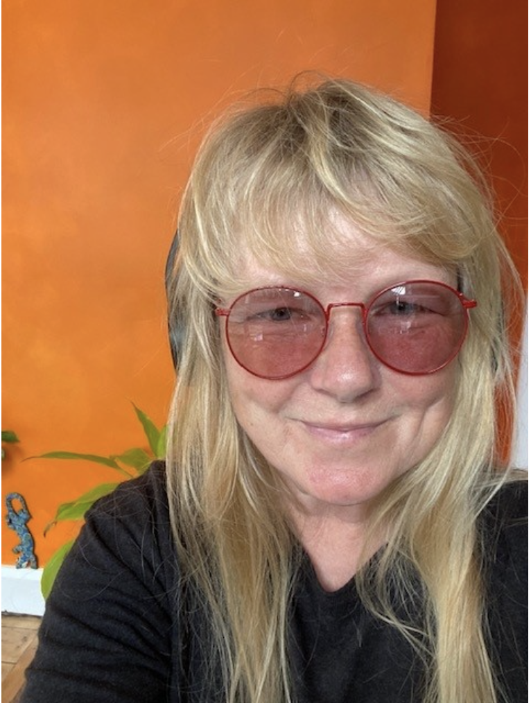 Ghizzi Dunlop smiles at the camera, she has pink tinted glasses with a bright orange wall behind her