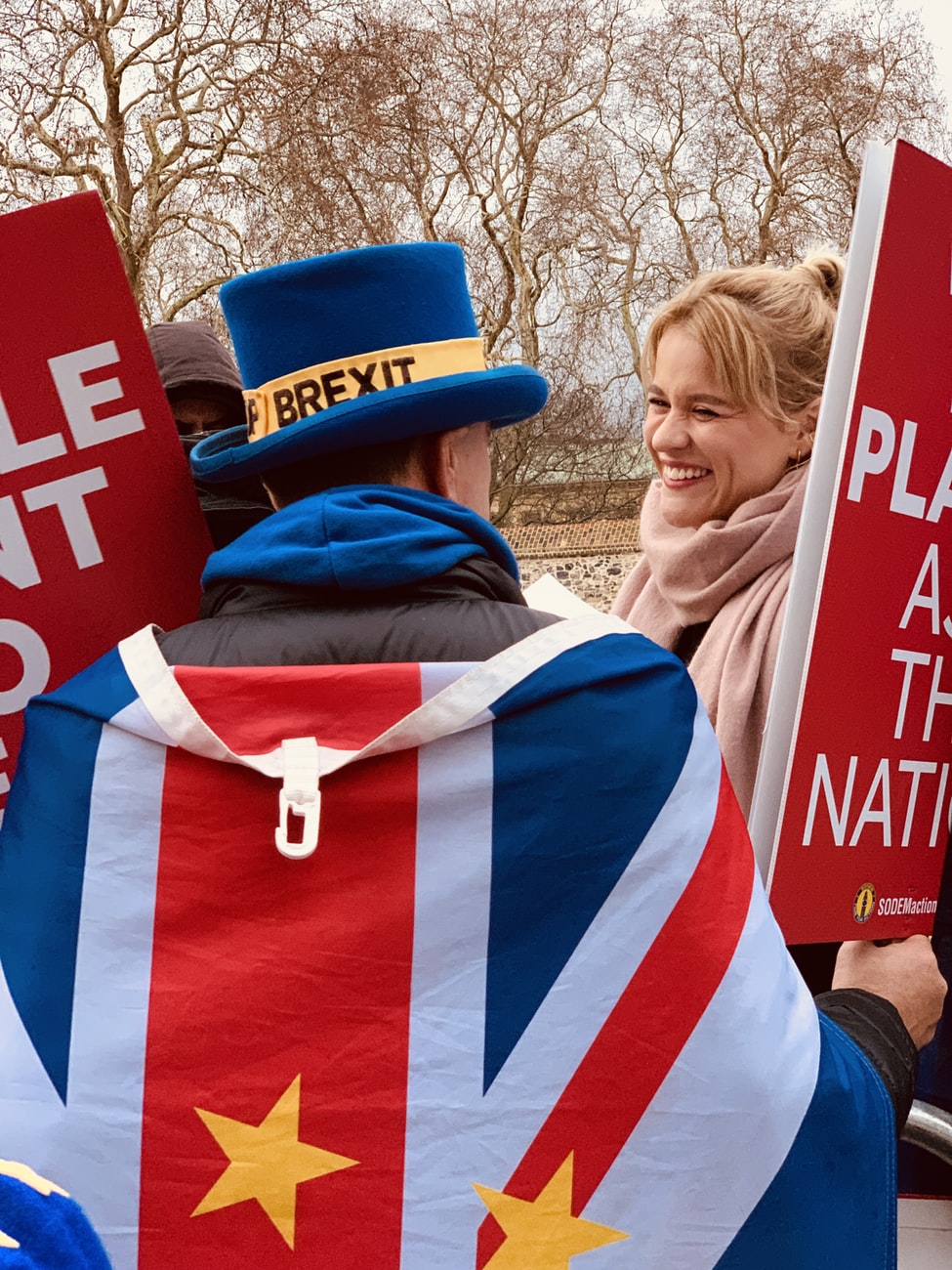 A picture of a man dressed in a top hat with Brexit on it draped in an EU flag. He is holding banners but text cannot be read