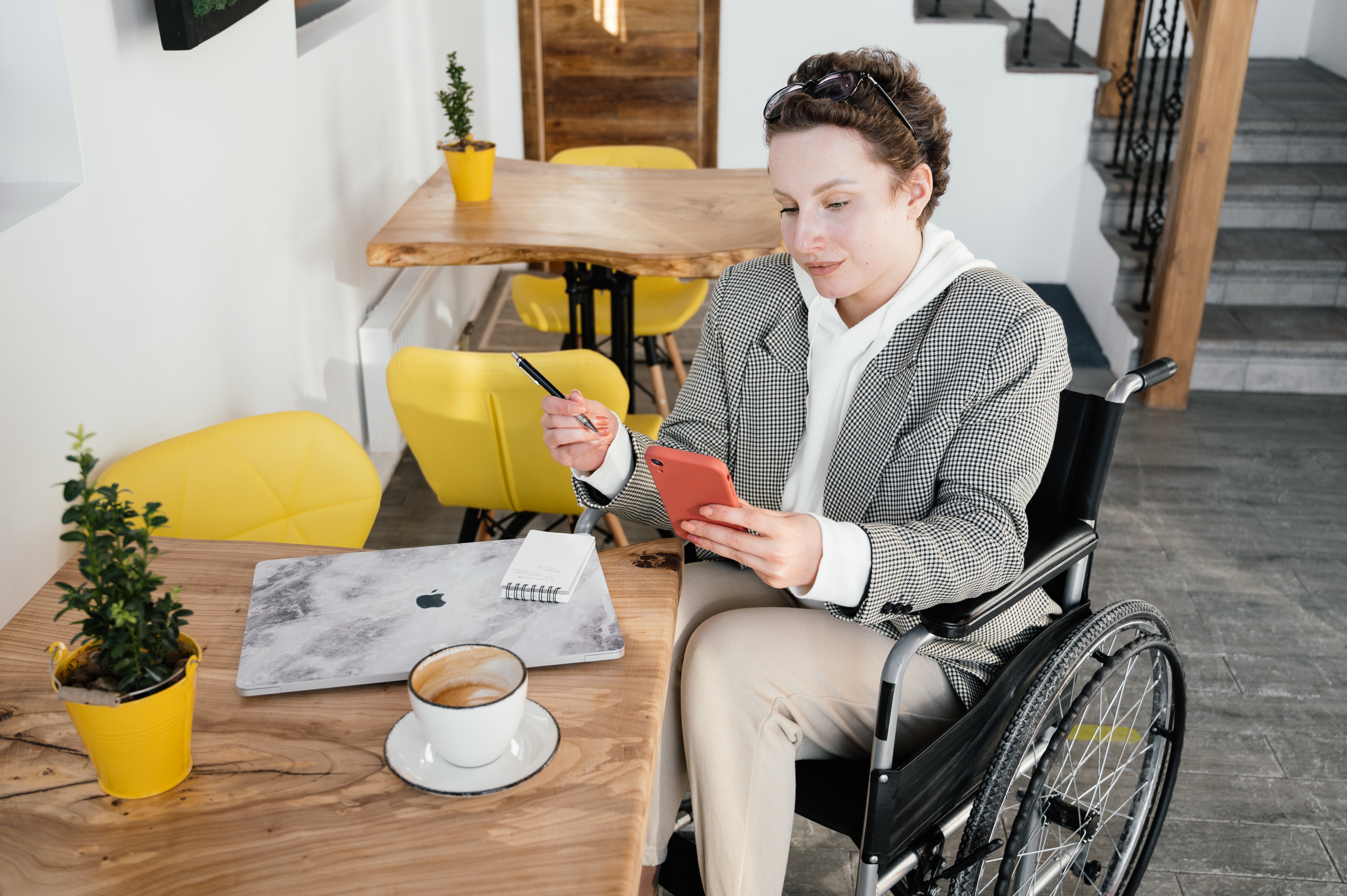 Image shows a woman in a wheelchair in a coffee shop. She in on her mobile pone and there is a laptop on the table
