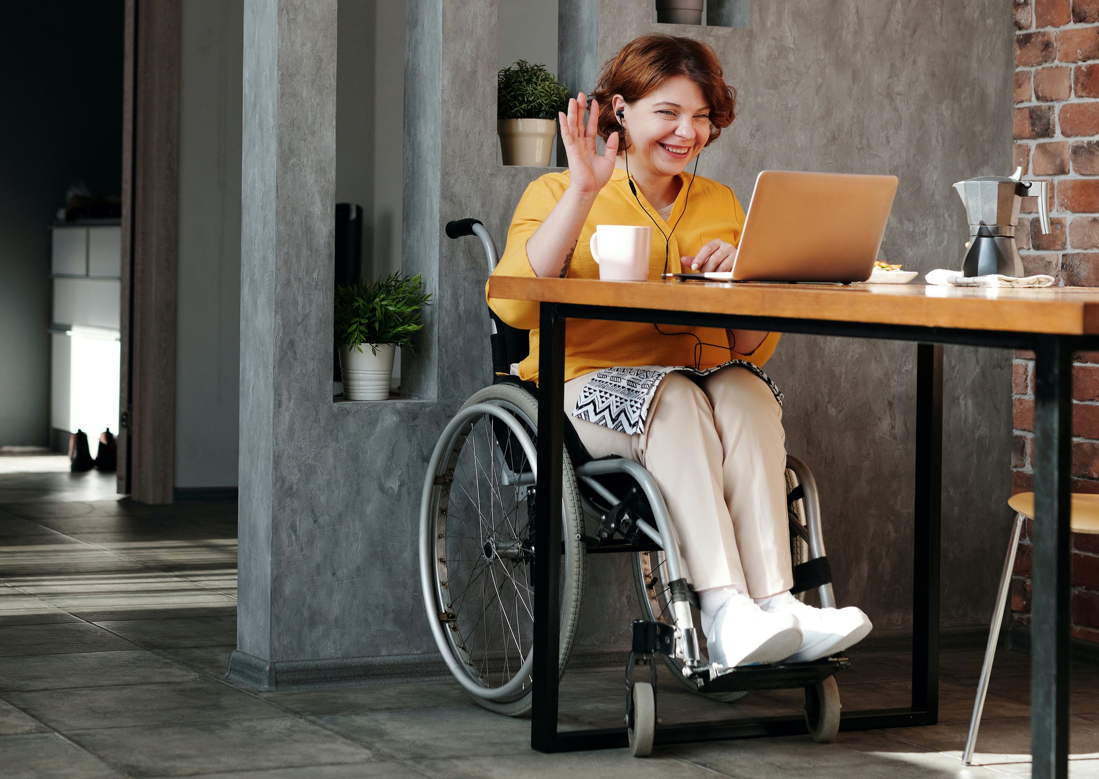 A woman in a wheelchair sitting at a table. She has a laptop and is smiling and waving at someone.