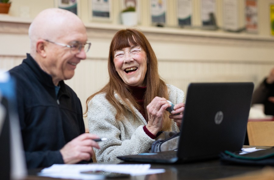 An older woman smiling sitting by a laptop in an office setting and older man sat next to her also smiling