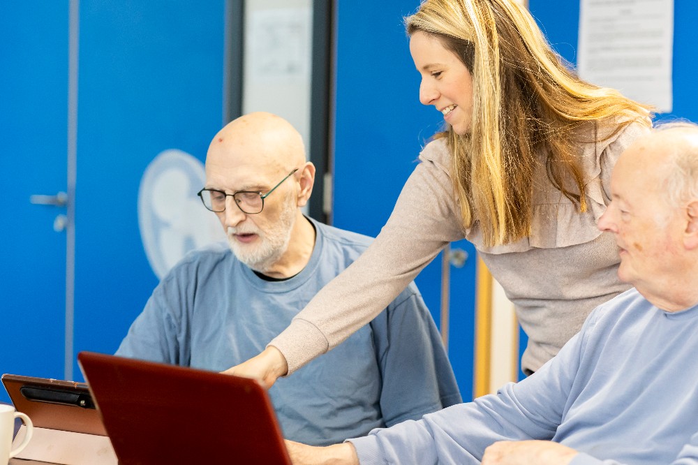 Two older gentlemen looking at laptops in learning setting with younger woman pointing at screen