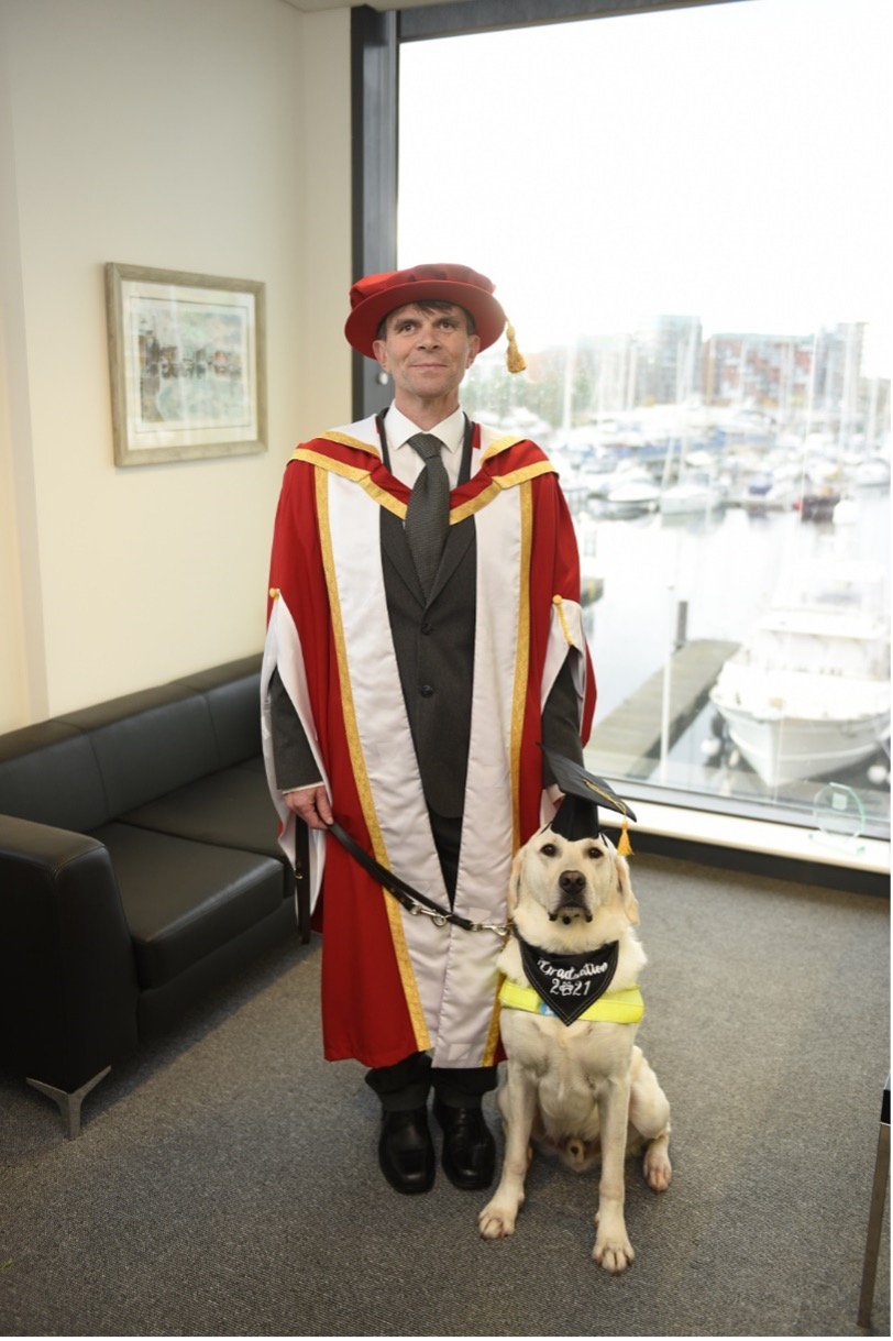 Robin Christopherson and guide dog Hugo receiving an honorary doctorate from the University of Suffolk Computer Science department